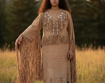 May include: A tan suede-like two-piece outfit with fringe and beaded details. The top has long sleeves with fringe, and the skirt has fringe and decorative elements. The outfit is worn in a field of tall grass.
