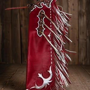 Peut inclure: Un pantalon de protection en cuir rouge profond orné de motifs floraux gravés sur la partie supérieure, d'accents argentés et de longues franges superposées en bandes rouges et blanches alternées le long de la jambe, avec une fine lanière.