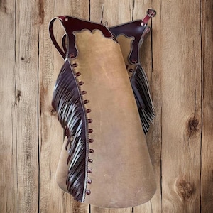 May include: Tan leather chaps with dark brown detailing and fringe. The chaps feature a scalloped edge at the top and rivets along the side. They are suspended against a wooden backdrop, showcasing a rustic aesthetic.
