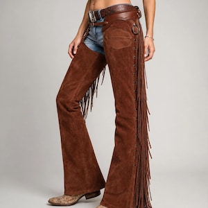 May include: Brown suede leather chaps featuring long fringe along the outer legs, a metal ring detail on the hip, and a decorative leather belt with a silver-toned buckle. The chaps are worn over blue denim jeans and paired with patterned boots.