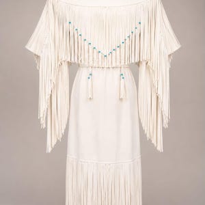 May include: An off-white dress featuring an extensive fringe overlay on the bodice and sleeves. The fringe is accented with small turquoise beads arranged in a V-shape. A matching thin belt with two hanging tassels and turquoise beads cinches the waist.