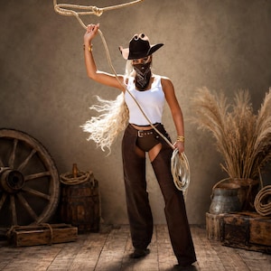 Puede incluir: Un sombrero de vaquero marrón oscuro, un pañuelo marrón estampado, una camiseta sin mangas blanca y chaparreras de cuero marrón oscuro sobre pantalones cortos marrones. Una cuerda enrollada se sostiene en la mano con un lazo hacia arriba.