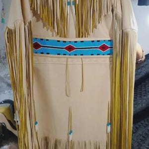 Könnte beinhalten: Ein beigefarbenes Kleid im indianischen Stil mit langen Ärmeln und Fransen. Das Kleid hat einen bestickten Gürtel mit roten, blauen und schwarzen Akzenten. Der Ausschnitt und die Ärmel sind mit Fransen und türkisfarbenen Perlen verziert.