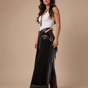 May include: Black suede chaps featuring a dark brown leather waistband with a buckle closure, silver-toned stud detailing along the sides, and long fringe trim. The chaps have a scalloped brown leather hem at the bottom and are worn over blue jeans.