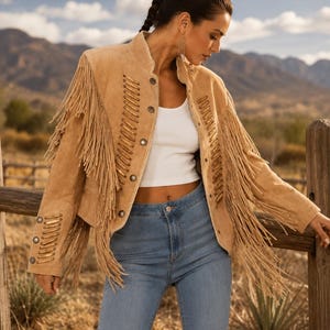 Puede incluir: Una chaqueta de ante color tostado con largos flecos en las mangas y paneles frontales. La chaqueta incluye un cuello alto, cierres de botones met&aacute;licos y detalles de cordones decorativos en la parte delantera y los pu&ntilde;os, sobre un top blanco y vaqueros azules.