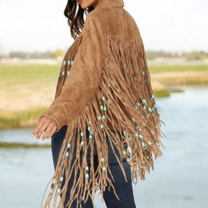 Puede incluir: Chaqueta de ante marrón con flecos largos y cuentas turquesas. La chaqueta tiene cuello y se lleva sobre unos vaqueros azul oscuro. Los flecos caen por la espalda y las mangas, creando un estilo bohemio.