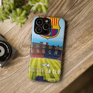 May include: A smartphone case featuring the FC Barcelona crest and a stadium scene. The case has a blue sky background and the words "ES QUE UN CLU" are visible. The case is on a wooden surface.