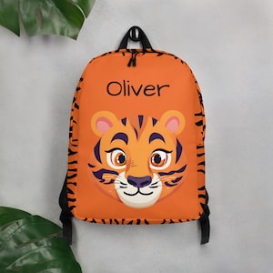 Op de afbeelding: Oranje rugzak met een tijgergezicht en zwarte strepen. De rugzak heeft een zwarte rits en de naam "Oliver" staat op de voorkant.