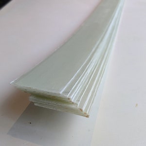 May include: A pile of thin, rectangular, pale green fiberglass strips. The strips are angled, showing their layered structure. The edges of the strips are a little rough. The background is a light, neutral colour.