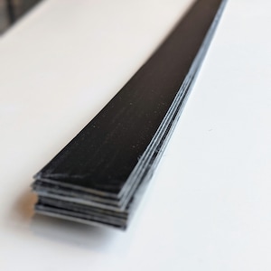 May include: A stack of thin, black rectangular pieces, possibly wood or composite material, are arranged on a white surface. The pieces are stacked neatly, with their edges aligned, creating a dark, linear form against the bright background.