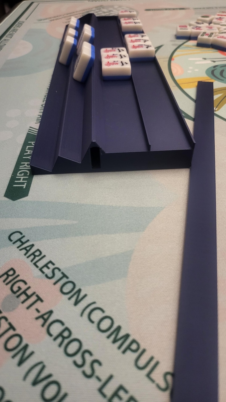 K&ouml;nnte beinhalten: Ein marineblauer Mahjong-Kartenhalter mit wei&szlig;en und blauen Karten. Die Karten haben rote und schwarze Markierungen. Das Spielbrett tr&auml;gt die Aufschrift "PLAY RIGHT" und "CHARLESTON (COMPULS".