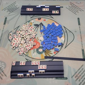 May include: A Mahjong game set with tiles, racks, and a game board. The tiles are white with black and red symbols. Blue racks hold tiles, and a pile of blue tiles sits in the center. The board has floral and text elements.