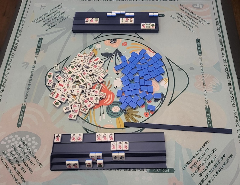 K&ouml;nnte beinhalten: Ein Mahjong-Spielset mit wei&szlig;en Spielsteinen mit schwarzen und roten Symbolen, angeordnet auf einem Brett mit Blumenmuster. Blaue Halterungen halten einige Spielsteine, mit einem Haufen blauer Spielsteine in der Mitte. Das Brett hat Text.