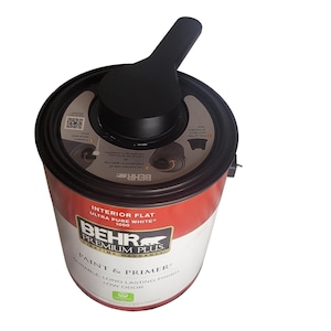 May include: A can of BEHR PREMIUM PLUS paint and primer in Ultra Pure White. The can has a black lid with a built-in handle and a red and white label. The paint can is designed for interior flat use.