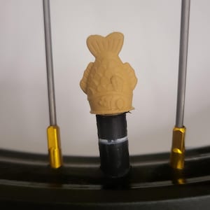 May include: A tan fish-shaped valve stem cap on a bicycle tire. The cap is detailed with scales and fins, and sits atop a black valve stem. The bicycle wheel has silver spokes and a black tire.