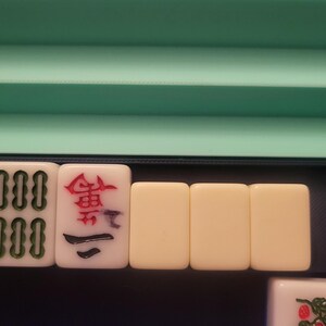 K&ouml;nnte beinhalten: Eine Auswahl an Mahjong-Steinen, darunter ein "Joker"-Stein mit gr&uuml;nem Design und rotem Text. Andere Steine zeigen verschiedene Symbole und Zahlen in Rot und Gr&uuml;n. Die Steine sind in einem Tablett mit gr&uuml;nem und blauem Hintergrund angeordnet.
