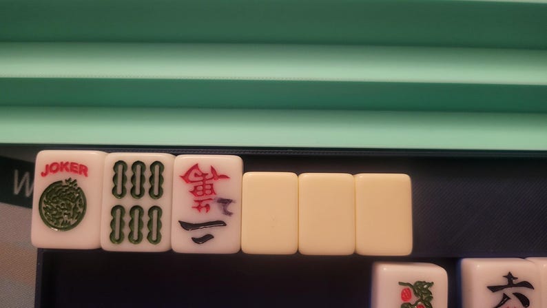 K&ouml;nnte beinhalten: Eine Auswahl an Mahjong-Steinen, darunter ein "Joker"-Stein mit gr&uuml;nem Design und rotem Text. Andere Steine zeigen verschiedene Symbole und Zahlen in Rot und Gr&uuml;n. Die Steine sind in einem Tablett mit gr&uuml;nem und blauem Hintergrund angeordnet.