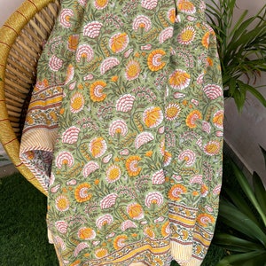 Op de afbeelding: Een groene katoenen deken met een bloemenprint in tinten roze, geel en groen. De deken heeft een rand met een gestreept patroon.