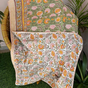 Op de afbeelding: Een groene en witte quilt met een bloemenpatroon en een gele en oranje bloemenrand. De quilt is gevouwen en tentoongesteld op een rieten stoel.