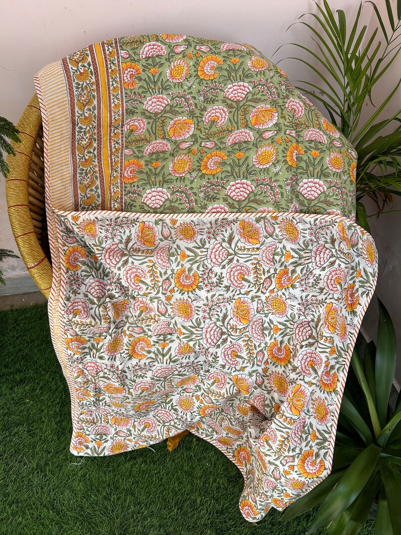 Op de afbeelding: Een groene en witte quilt met een bloemenpatroon en een gele en oranje bloemenrand. De quilt is gevouwen en tentoongesteld op een rieten stoel.
