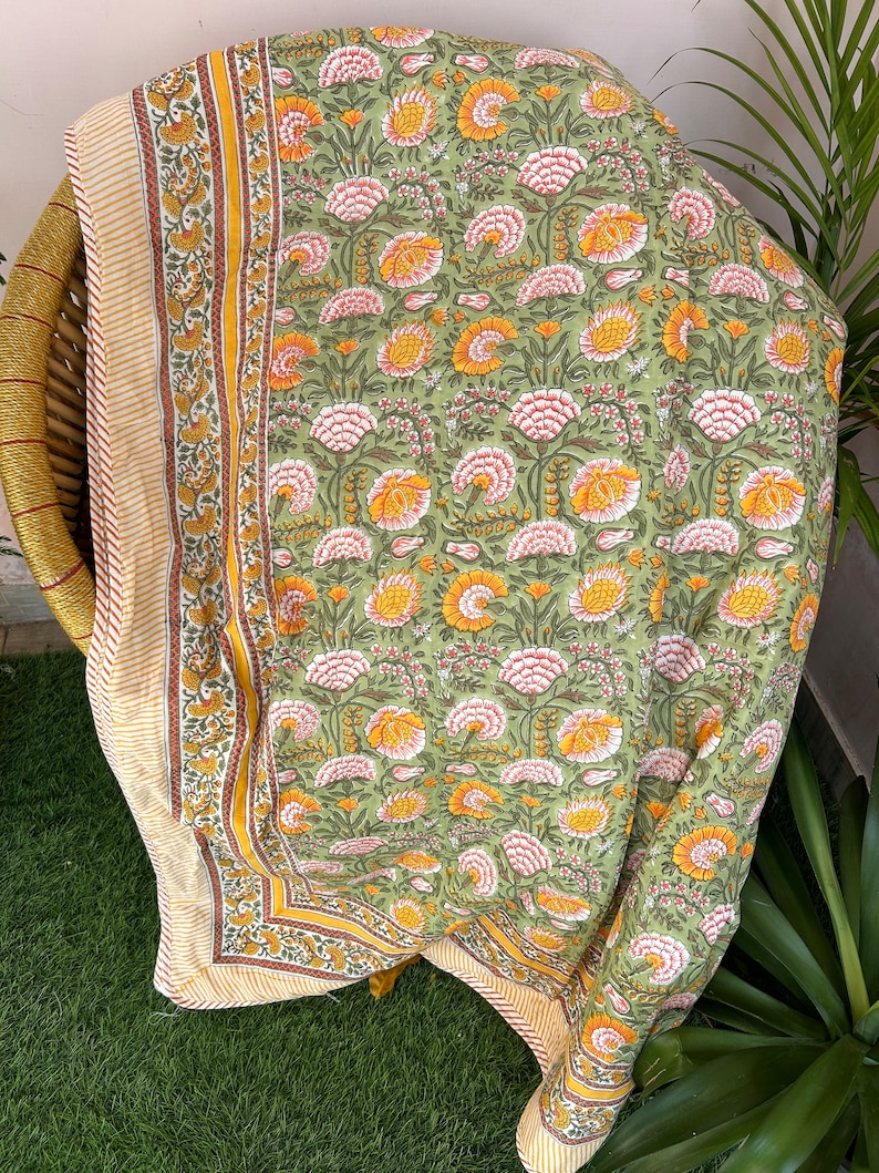 Op de afbeelding: Een groene en gele katoenen deken met een bloemenpatroon en een gele en oranje rand. De deken is gevouwen en wordt getoond op een rieten stoel.
