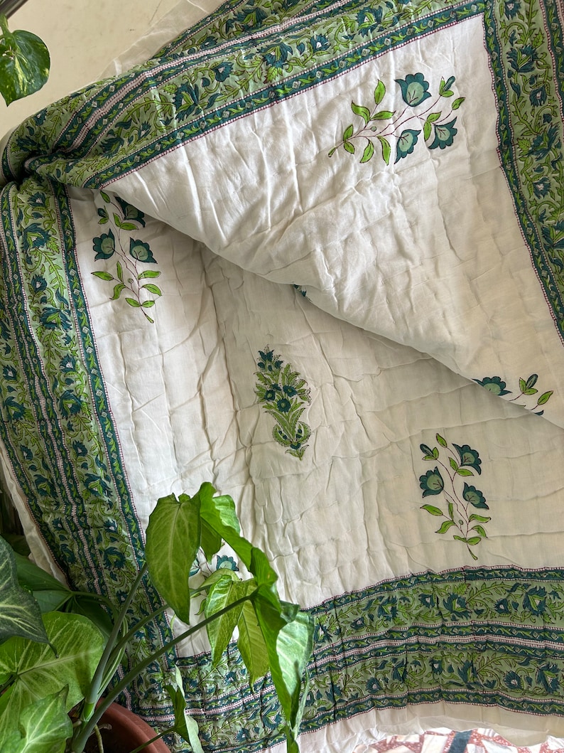 May include: White cotton quilt with a green floral print. The quilt has a border of green and white floral designs. The quilt is folded in half and the floral designs are visible on both sides.
