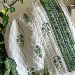 May include: White cotton quilt with a green floral print and a green and white striped border. The quilt is folded and hanging on a metal railing.