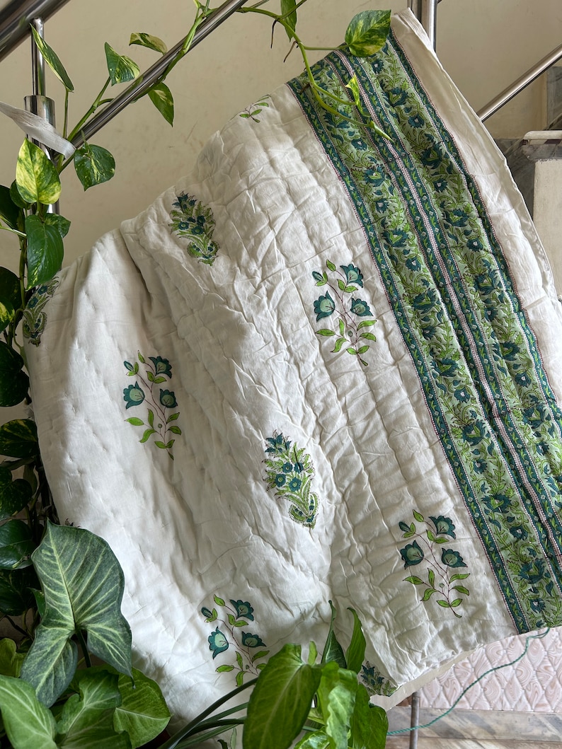 May include: White cotton quilt with a green floral print and a green and white striped border. The quilt is folded and hanging on a metal railing.