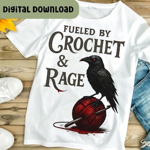 Puede incluir: Camiseta blanca con el texto "Fueled by Crochet & Rage" y un gráfico de un cuervo negro con ojos rojos posado sobre una bola de hilo roja. Un ganchillo está insertado en el hilo. "Digital Download" en la parte superior izquierda.