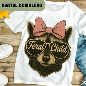 以下が含まれることがあります： 「Feral Child」の文字が入ったサングラスをかけたアライグマが描かれた白いTシャツ。アライグマはピンクのリボンをしています。「DIGITAL DOWNLOAD」の文字が画像上部にあります。