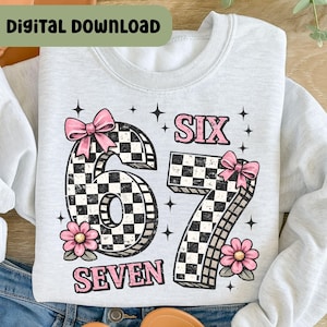 May include: A light gray sweatshirt with the words "SIX SEVEN" in pink, and the numbers "67" in a black and white checkered pattern. The sweatshirt has pink bows and flowers.
