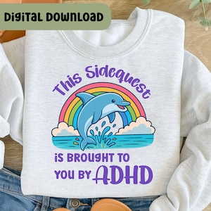 Puede incluir: Sudadera gris claro con un diseño de un delfín azul saltando del agua frente a un arcoíris. El texto dice "This Sidequest is Brought to You by ADHD" en morado y azul.