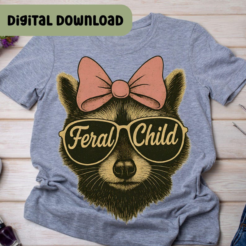 以下が含まれることがあります： 「Feral Child」の文字が入ったサングラスとピンクのリボンをつけたアライグマが描かれたグレーのTシャツ。「Digital Download」の文字がTシャツの上部にあります。