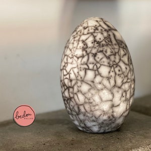 May include: A white ceramic egg with a crackled glaze and black lines. The egg is sitting on a gray surface.