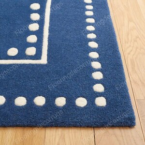 May include: A section of a blue rug with a white dotted border and a white square design in the corner. The rug is made of a soft, textured material and is placed on a light-colored wooden floor.