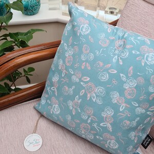 May include: A teal blue cushion with a white and pink floral pattern. The cushion is sitting on a light pink chair with a wooden frame.