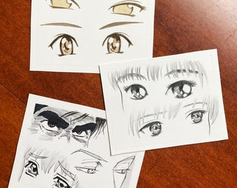 May include: Three white rectangular cards featuring black ink and marker illustrations of various anime-style eyes. The drawings depict diverse eye shapes, expressions, and brow styles, including detailed irises, lashes, and shading.