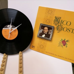 Puede incluir: Un disco de vinilo negro con una esfera de reloj y agujas blancas. El disco está montado en un soporte de madera. El disco está al lado de un álbum de disco amarillo con el título "Nico Dostal" y una foto de un hombre con esmoquin. El texto del álbum dice "Das Schönste aus Operette und Tonfilm".