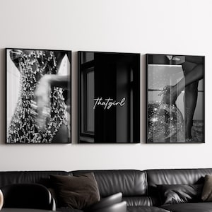 May include: A triptych of framed black and white art prints. The central print features the text "thatgirl". The other two prints display abstract imagery. The artwork hangs above a black leather sofa in a modern living room setting.