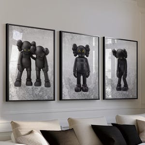 May include: Three framed black and white art prints featuring cartoon-like figures with X-shaped eyes. The prints hang above a white sectional sofa with black and neutral-colored throw pillows. A wooden side table and a black coffee table complete the living room.