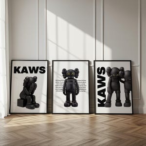 May include: Three framed art prints featuring black KAWS figures. The prints have white backgrounds and black frames. The KAWS figures have black bodies with X-shaped eyes. The word "KAWS" is printed in large, bold, black letters on two of the prints.