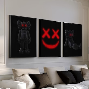 K&ouml;nnte beinhalten: Drei gerahmte schwarze Kunstdrucke mit roten Akzenten. Die Drucke zeigen eine stehende Figur, ein l&auml;chelndes Gesicht und eine sitzende Figur. Die Drucke werden &uuml;ber einem wei&szlig;en Ecksofa mit schwarzen und beigen Kissen angezeigt.