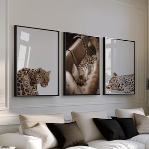 May include: Three framed art prints with leopard images adorn a living room. The prints, in black frames, show leopards in different poses. The room has a neutral colour scheme, a white sectional sofa, and a wooden side table. The sofa has black cushions.