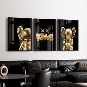 May include: Set of three framed art prints featuring gold-colored KAWS figures against a black background. The middle print displays the KAWS logo. The artwork is displayed above a black leather sofa.