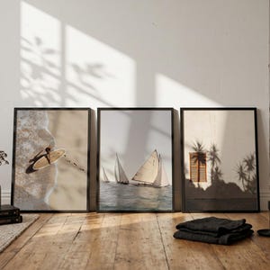 Op de afbeelding: Drie ingelijste wandposters op een rij. De linker poster toont een luchtfoto van een surfplank op een zandstrand. De middelste poster toont zeilboten op het water. De rechter poster toont een muur met een raam en schaduwen van palmbomen.