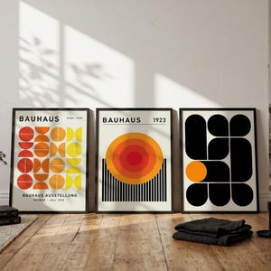 May include: Three framed art prints featuring Bauhaus-inspired graphic designs. The left print displays orange and yellow geometric semicircles with text reading BAUHAUS AUSSTELLUNG WEIMAR JULI 1923. The center print shows a large orange circle above black lines.