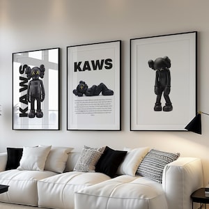 May include: Three framed KAWS art prints hang above a white sectional sofa. The prints feature black figures with the word "KAWS" in bold letters. The living room has a neutral color palette with a black coffee table and a gray rug.