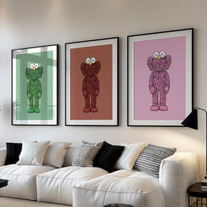 May include: Three framed art prints featuring cartoon figures in green, brown, and pink, each with an X-shaped eye design. The prints hang above a white sectional sofa with black and patterned throw pillows. A black coffee table and a floor lamp are also visible.