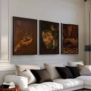May include: Three framed art prints with a vintage aesthetic. One shows a hand holding a crown, another features lipstick kisses on a tray, and the third depicts a person playing chess. The prints are displayed above a white sectional sofa.