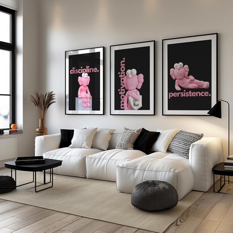 May include: Three framed art prints featuring pink cartoon figures with the words "discipline," "motivation," and "persistence" in pink text on a black background. The prints hang above a white sectional sofa.